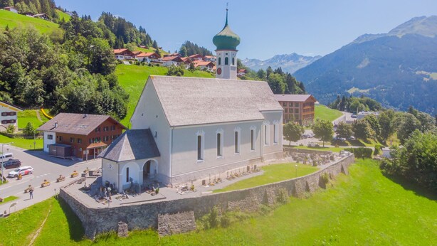 A church with a bell tower, surrounded by a stone wall and a small courtyard.