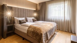 A modern bedroom with a large bed, a headboard with built-in lighting, a bedside table, and a window with curtains.