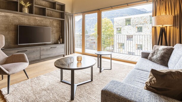 A modern living room with a flat-screen TV, a sofa, a coffee table, and a balcony with a view of buildings and mountains.