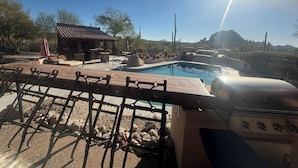 A pool area with a barbecue grill, outdoor seating, and a small building with a tiled roof.