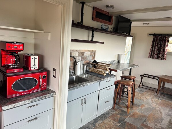 Una cocina compacta con microondas rojo, tostadora y cafetera sobre la encimera, un fregadero y un taburete de madera.