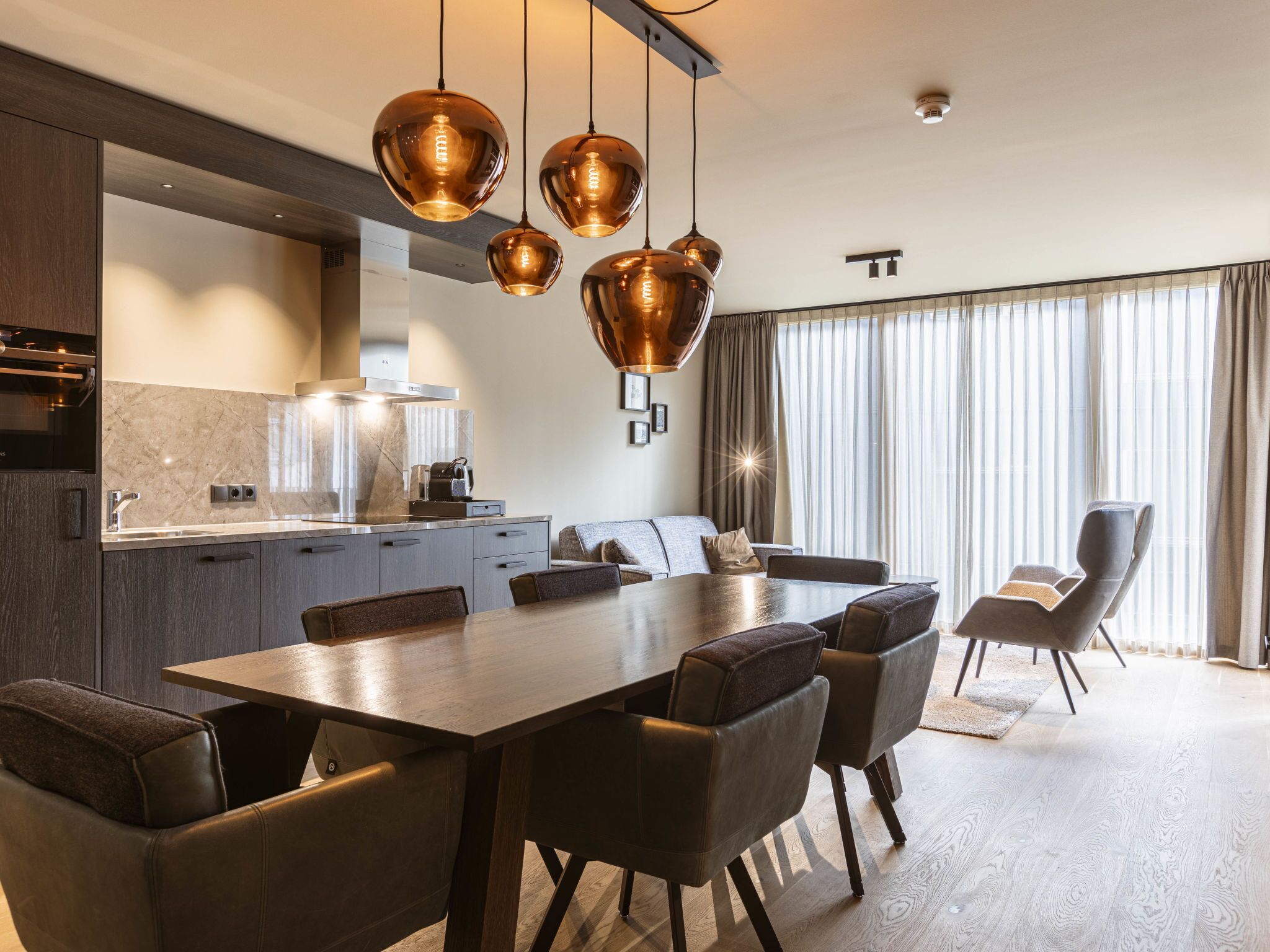 Eine moderne Küche mit einer zentralen Kochinsel, einem Essbereich mit großem Tisch und Stühlen sowie einem Wohnbereich mit Sofa und Relaxsessel.
