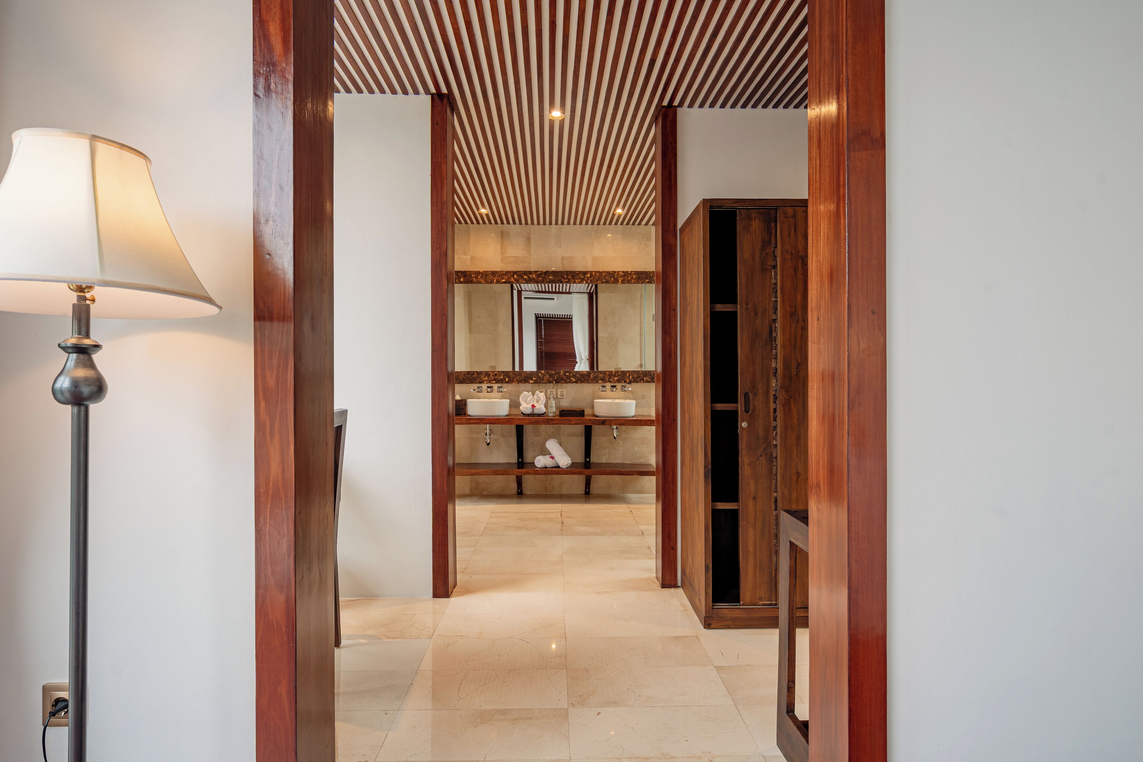 A hallway leading to a bathroom with a wooden door, a mirror, and a floor lamp.