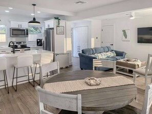 A modern kitchen with white cabinets, a dining area with a round table, and a blue sofa in a bright living room. - Surf Chic 3br, Firepit, Boating, Fishing, Beaches (Melbourne)