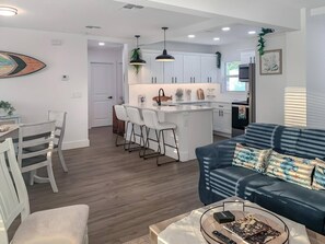 A modern kitchen with white cabinets, a central island, and bar stools. There is a blue sofa with a turtle pillow in the living area. - Surf Chic 3br, Firepit, Boating, Fishing, Beaches (Melbourne)