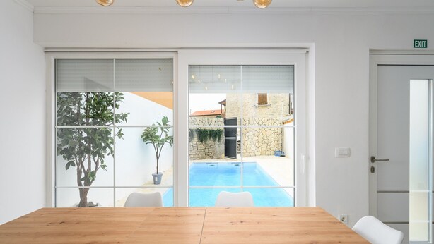 A conference room with a wooden table, white chairs, a glass partition, and a view of a swimming pool and stone building. - Villa in Porec With Private Pool and Beach (Porec)