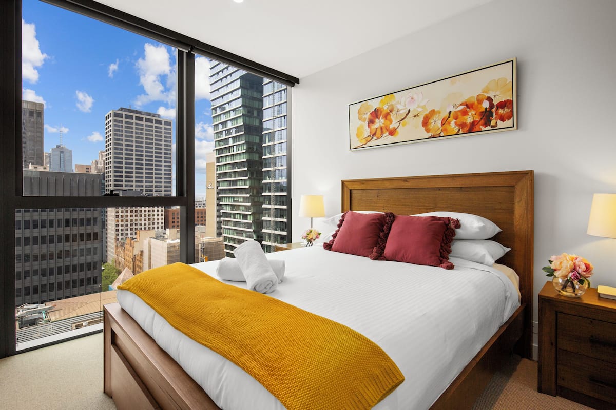 Ein Hotelzimmer mit großem Fenster und Blick auf die Stadt, einem ordentlich bezogenen Bett mit gelber Decke und einem Blumenbild an der Wand.