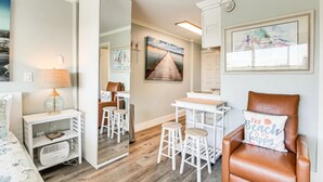 A modern, well-lit room with a wooden floor, a white desk with two stools, a brown leather chair, and a large mirror. - 114 Beachfront Heated Pool Studio Harborside View (Galveston)