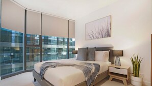 A modern bedroom with a large bed, a bedside table with a lamp, a potted plant, and a city view through floor-to-ceiling windows. - Melbourne Corporate Apartments – Collins Street (Melbourne)
