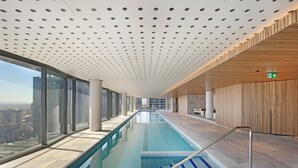 A modern indoor swimming pool with a view of the cityscape through large windows. - Melbourne Corporate Apartments – Collins Street (Melbourne)