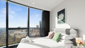 Een moderne slaapkamer met een groot raam, een bed met witte lakens, een grijs hoofdeinde en een nachtkastje met een bloevas.