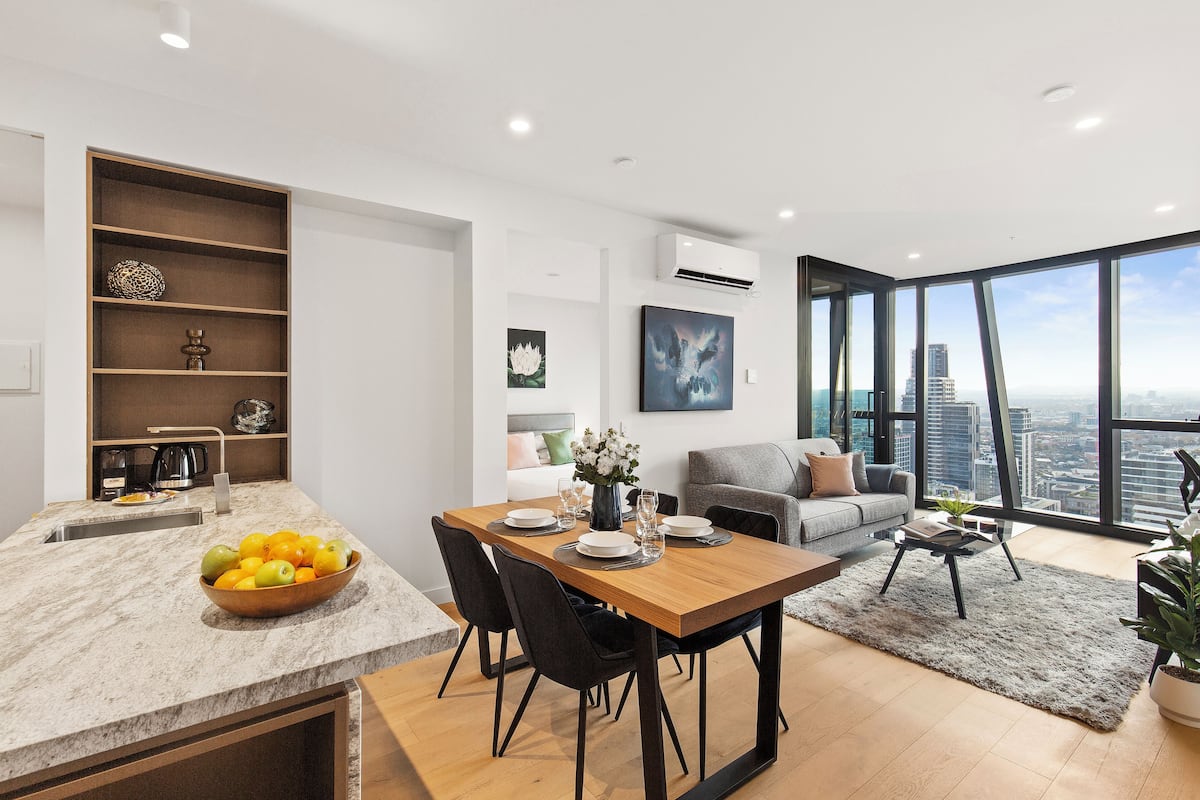 Una cocina moderna con encimera de mármol, comedor con mesa de madera y sillas negras, sofá gris y vistas a la ciudad a través de grandes ventanales.