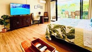 Quarto de hotel com cama, televisão, ventoinha no teto, um banco de madeira, uma mesinha e vista para o verde através de grandes janelas.