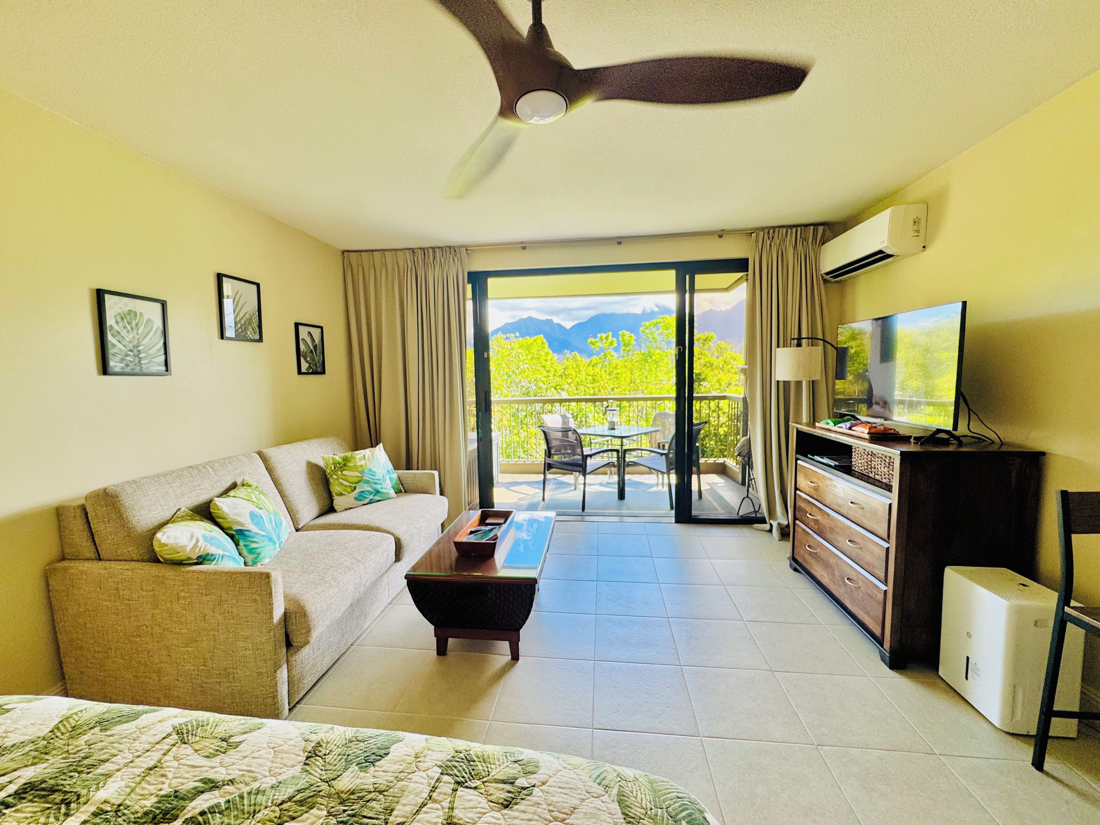 A hotel room with a bed, a sofa, a TV, a desk, a ceiling fan, and a balcony with a view of greenery and mountains.