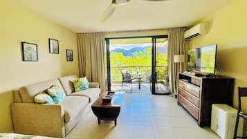 A hotel room with a bed, a sofa, a TV, a desk, a ceiling fan, and a balcony with a view of greenery and mountains.