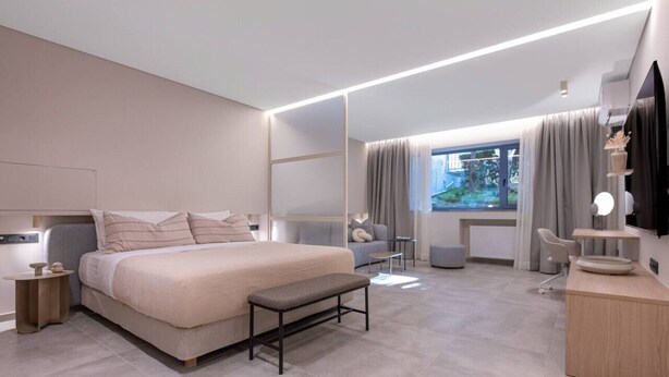 Une chambre moderne avec un grand lit, des tables de chevet, un banc, un bureau et une vue sur l’extérieur.