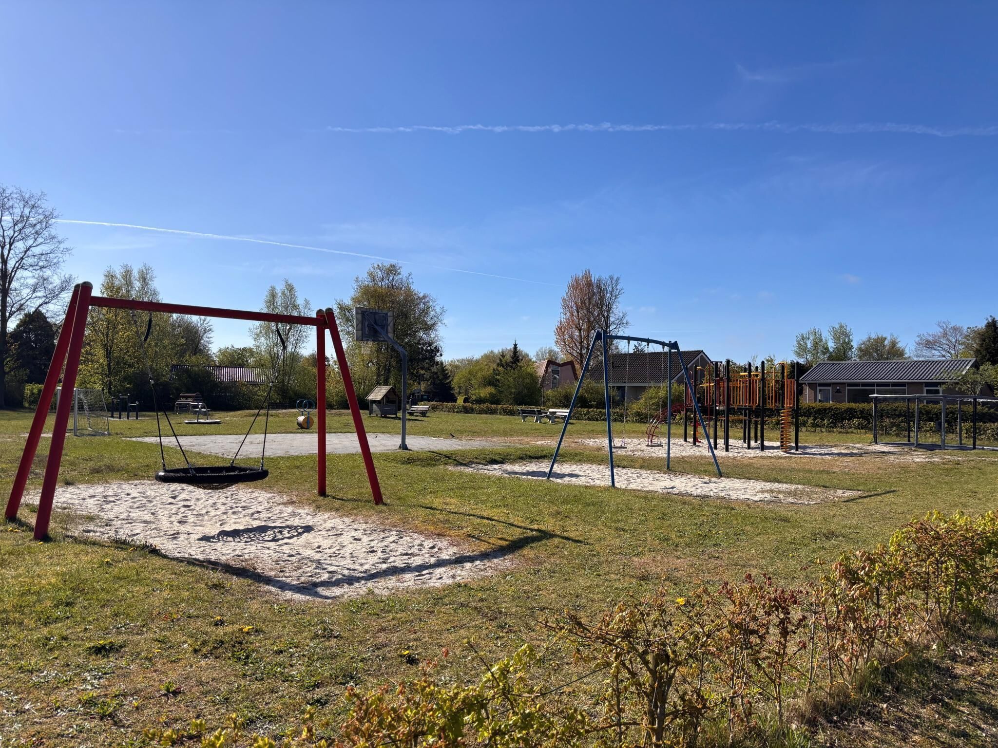 A playground with swings and climbing structures, surrounded by grass and trees.