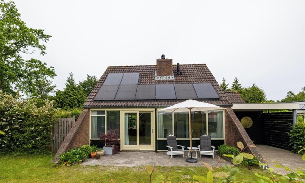 Uma casa com painéis solares, porta de entrada em vidro, um pátio com cadeiras e uma placa com o número 117.