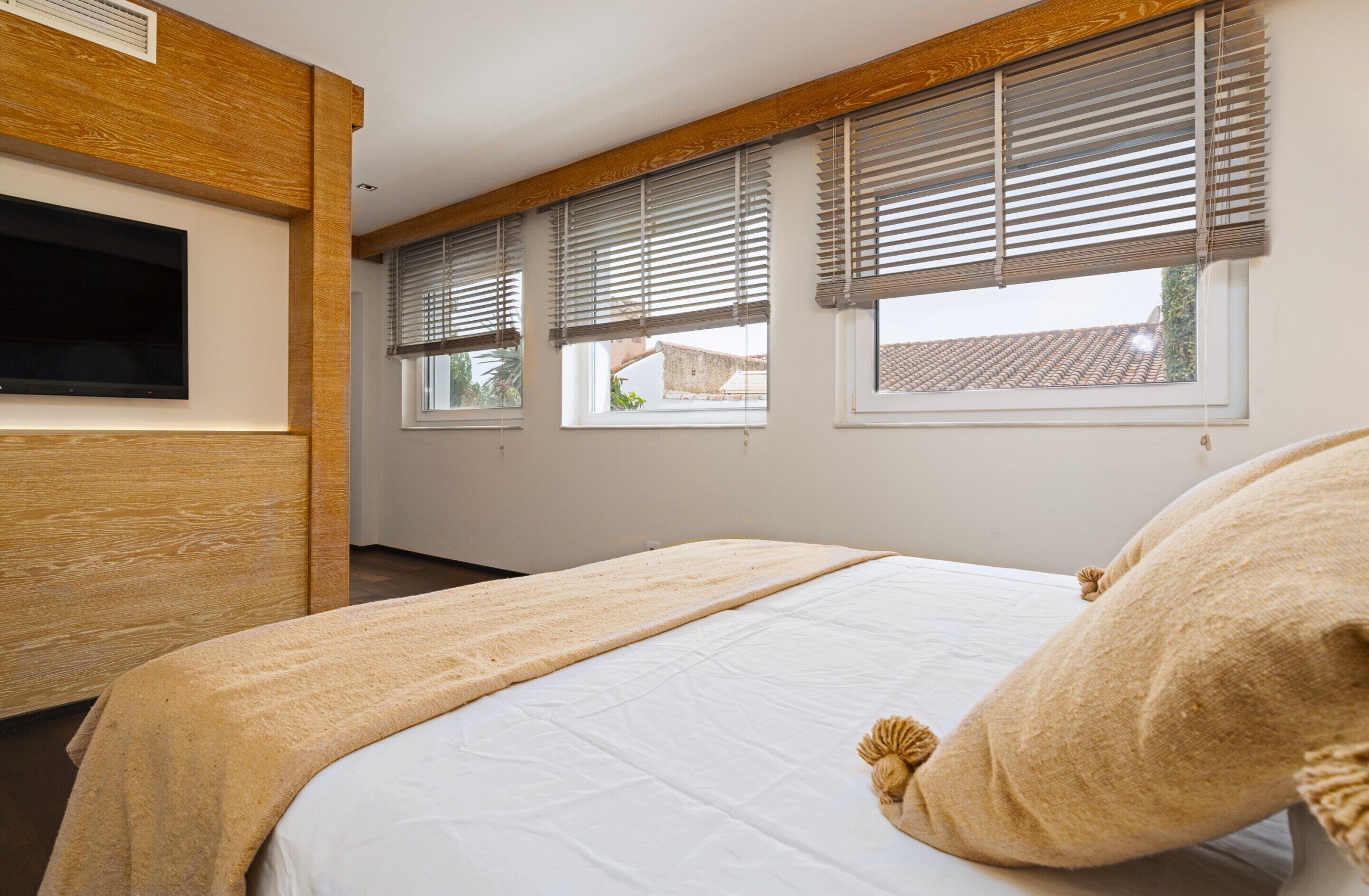 A bedroom with a bed, wooden headboard, a flat-screen TV, and three windows with blinds.