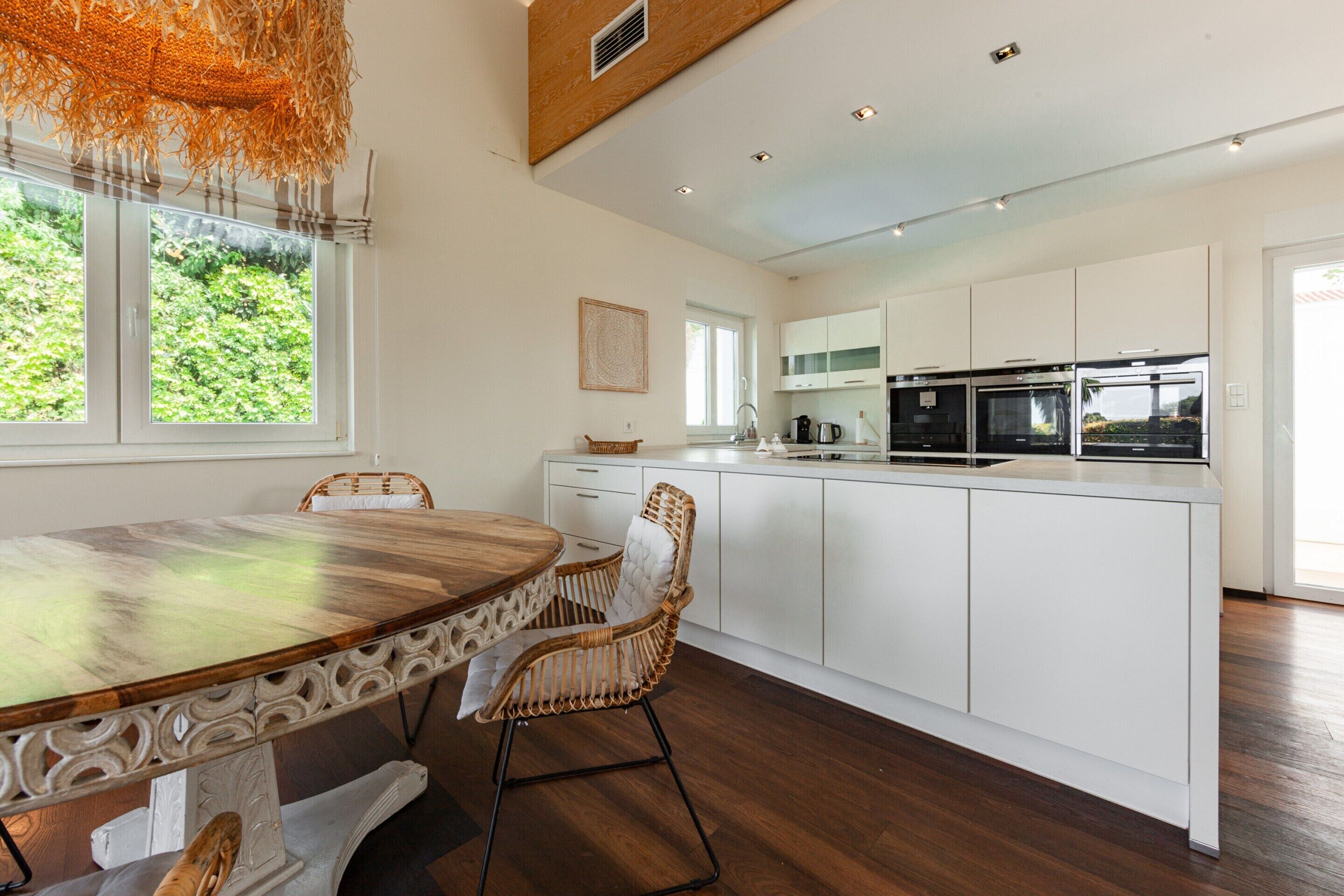 A modern kitchen with white cabinetry, a central island, and a dining area with a wooden table and wicker chairs.
