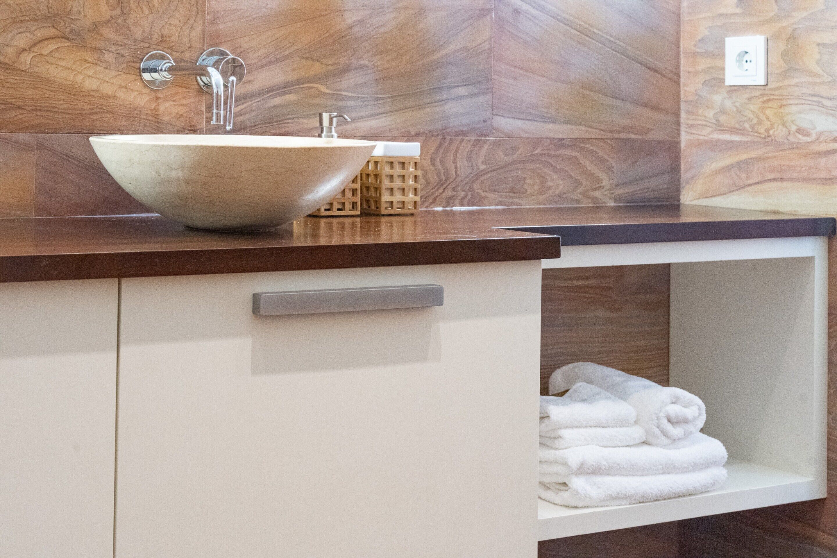 A bathroom with a large, round, beige sink on a dark countertop, a towel rack with folded towels, and a wooden wall with a marble-like pattern.