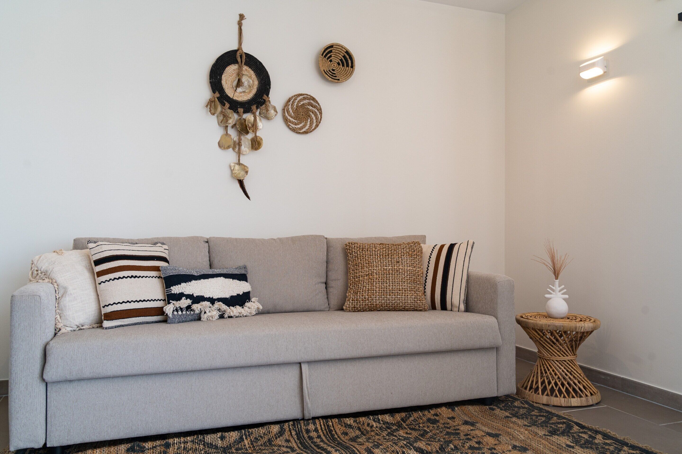 Un salon aux couleurs neutres, avec un canapé, des coussins à motifs, une décoration murale tissée et une petite table d’appoint sur laquelle est posé un vase.