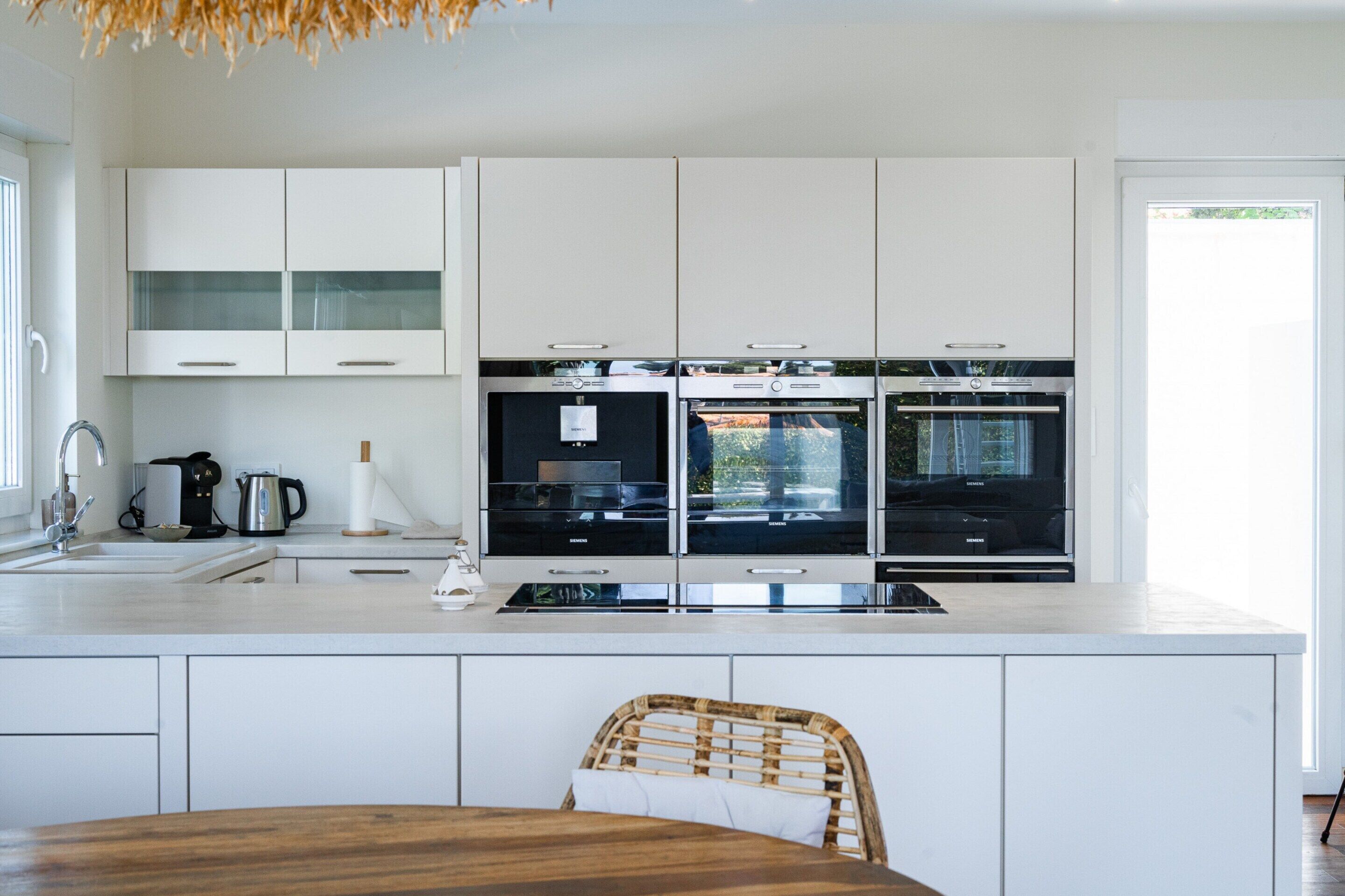 Una cucina moderna con mobili bianchi, forno integrato e zona pranzo con tavolo in legno e sedia in vimini.