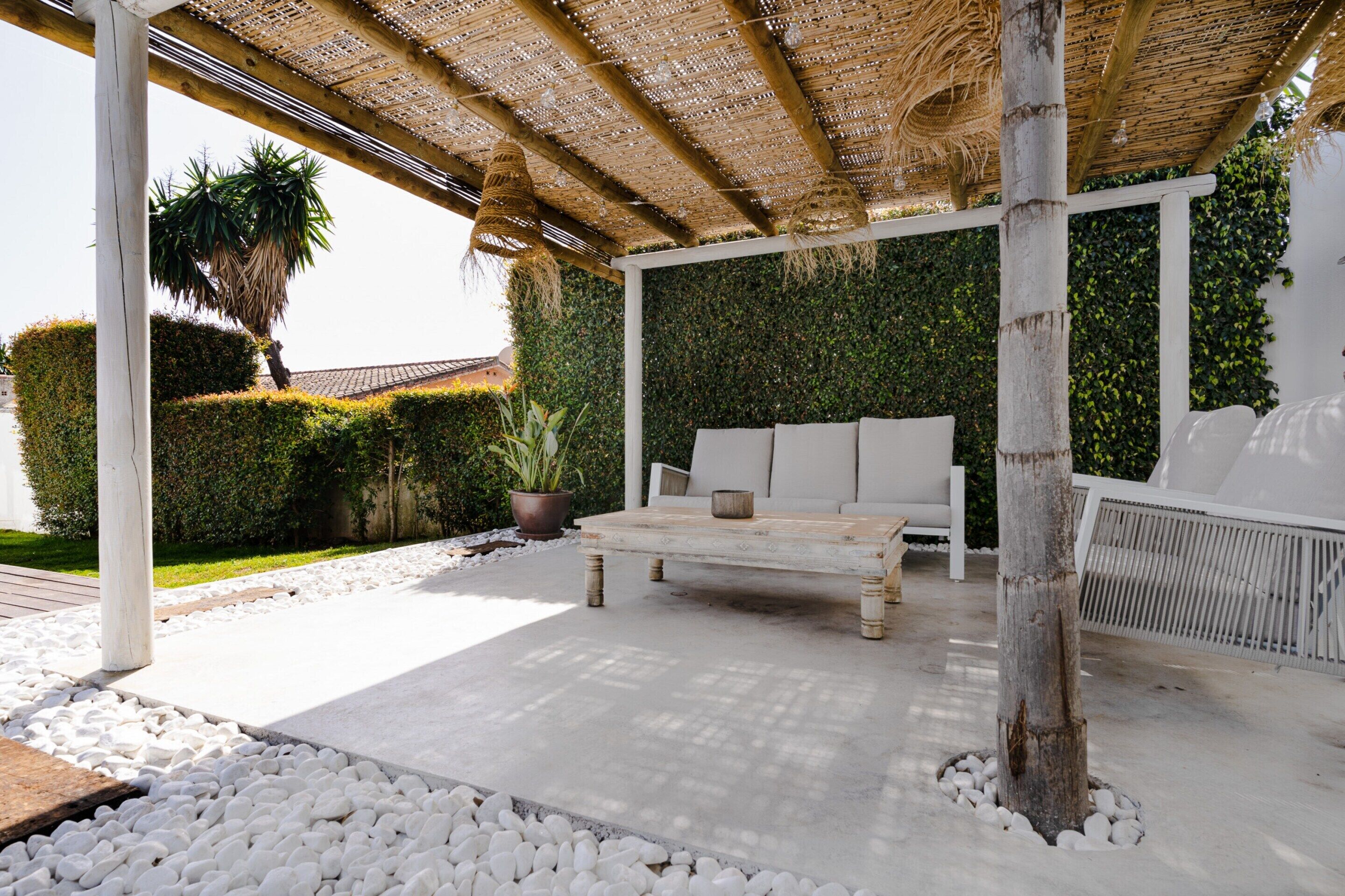 A covered outdoor seating area with a white sofa and a wooden table, surrounded by white pebbles and greenery.