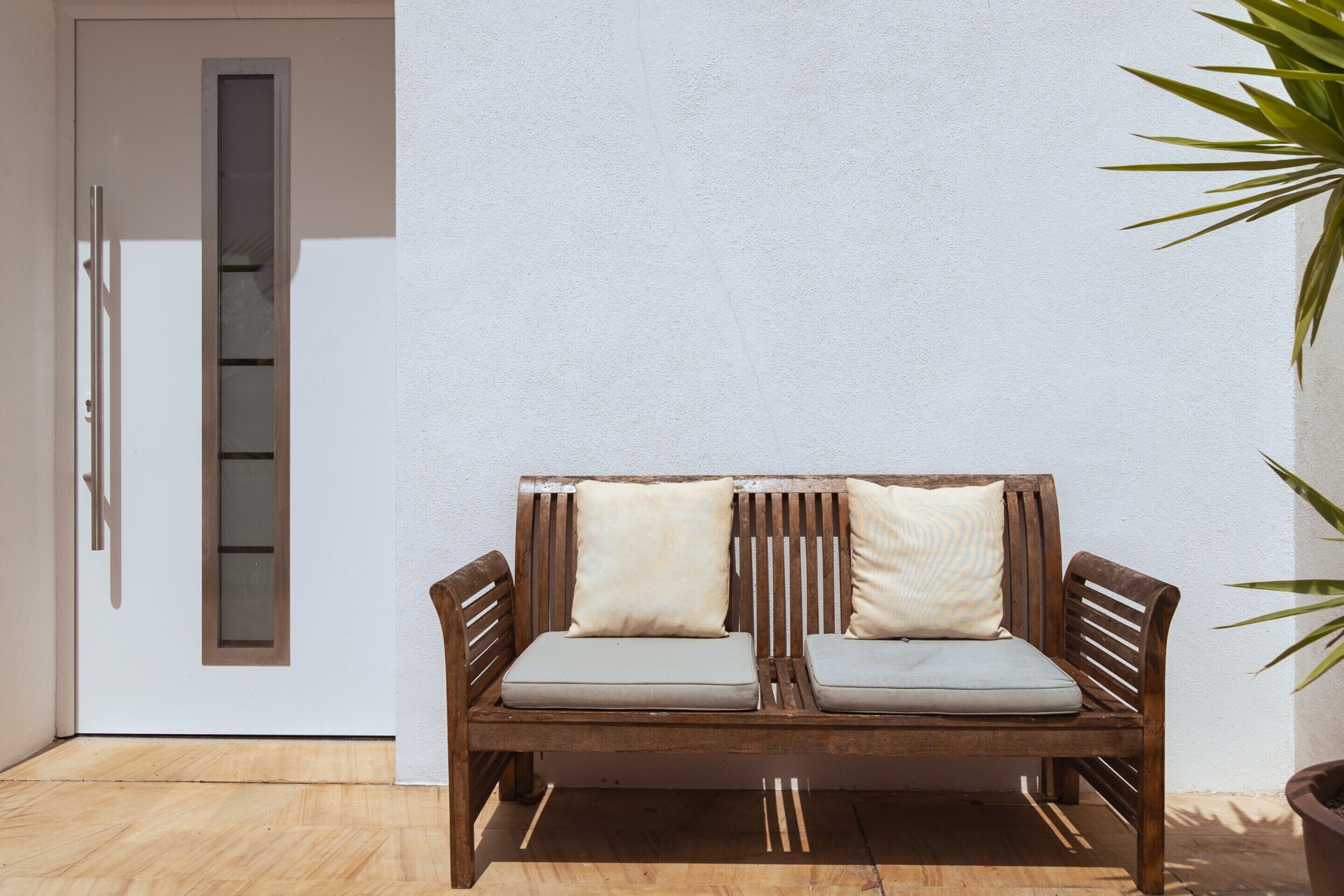 A wooden bench with cushions against a white wall, a door with a window, and a potted plant.