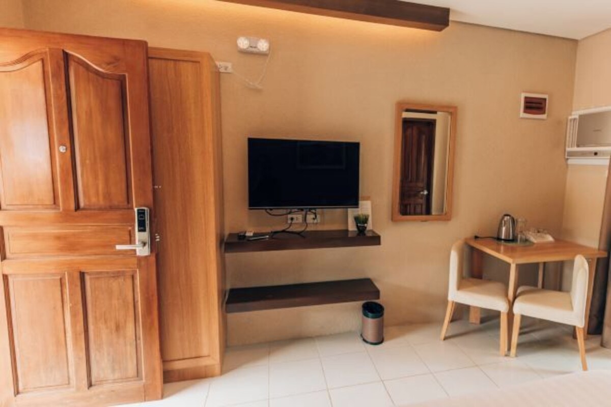 A compact room with a wooden wardrobe, a television on a stand, a small table with chairs, and a trash bin.