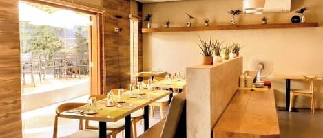 A restaurant with wooden tables and chairs, a long counter, and potted plants on shelves.