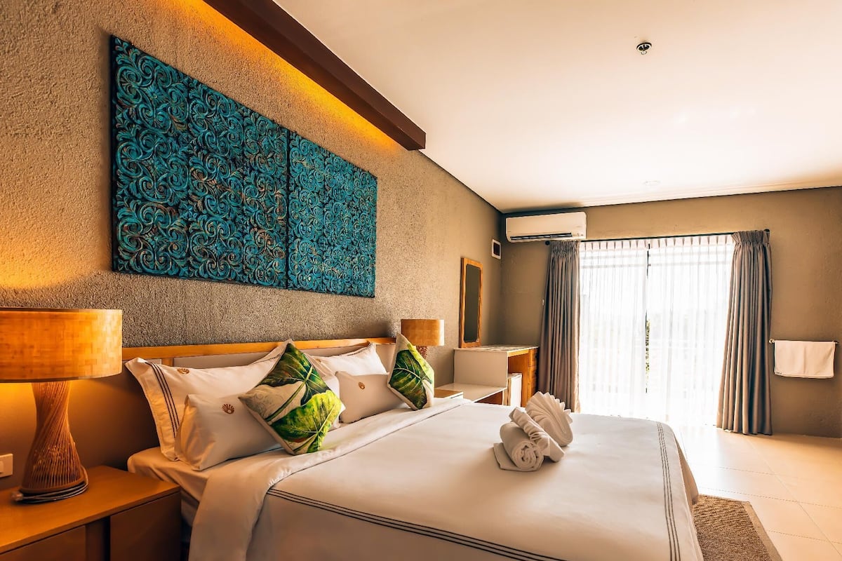 Quarto de hotel com uma cama grande, duas mesas de cabeceira com abajures, uma escrivaninha, um espelho e uma janela com cortinas.