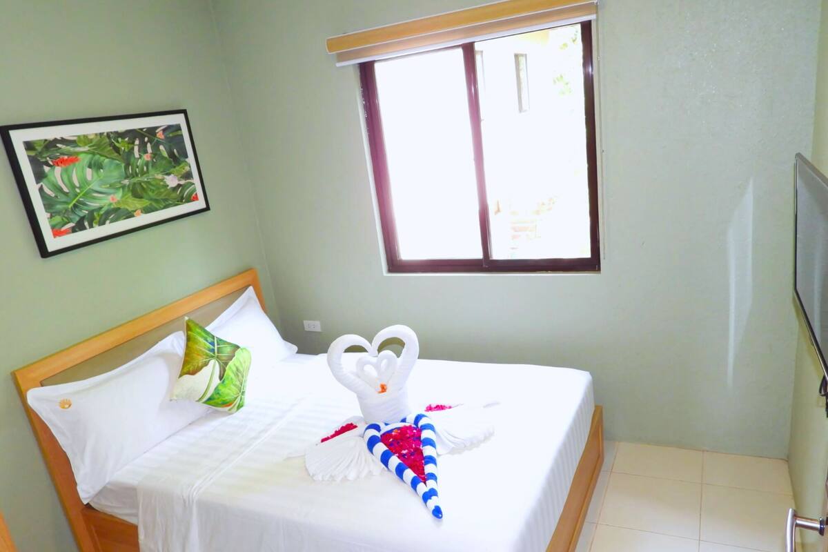 Cama bem arrumada com lençóis brancos, toalha em formato de coração e manta listrada. Há um travesseiro verde, uma moldura na parede e uma janela com persianas.