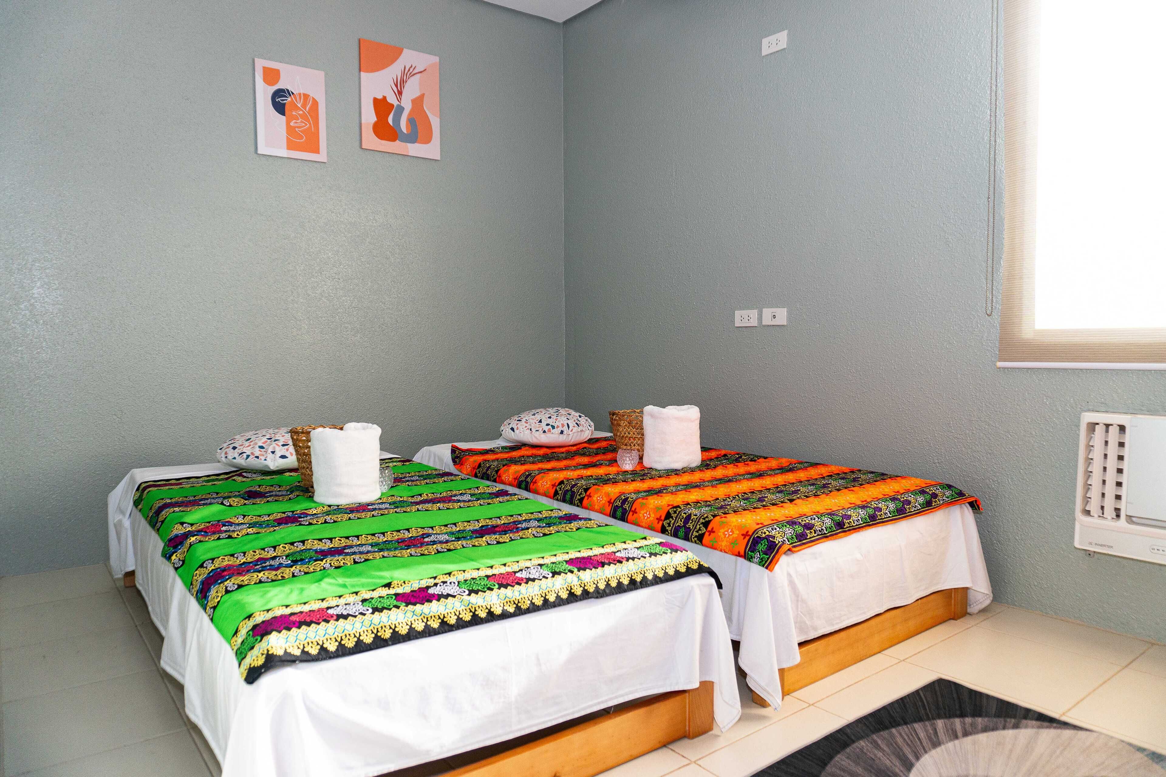 A room with two beds, each with colorful bedding and pillows, a window with blinds, and a wall with two framed pictures.