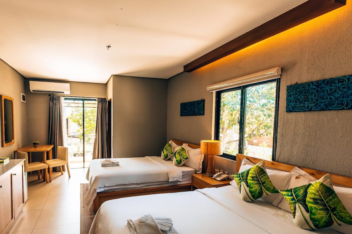 Quarto de hotel com duas camas, uma escrivaninha e uma janela com vista para o verde.