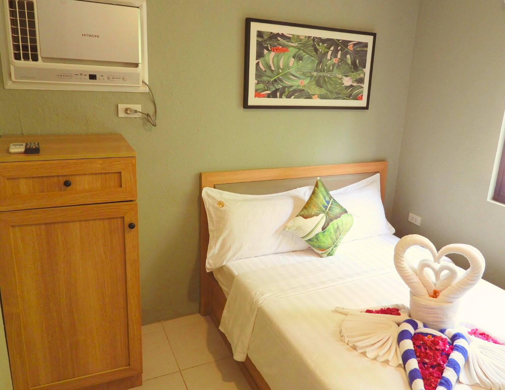 A hotel room with a bed, a wooden nightstand with a remote control, a wall-mounted air conditioner, and a framed tropical artwork.