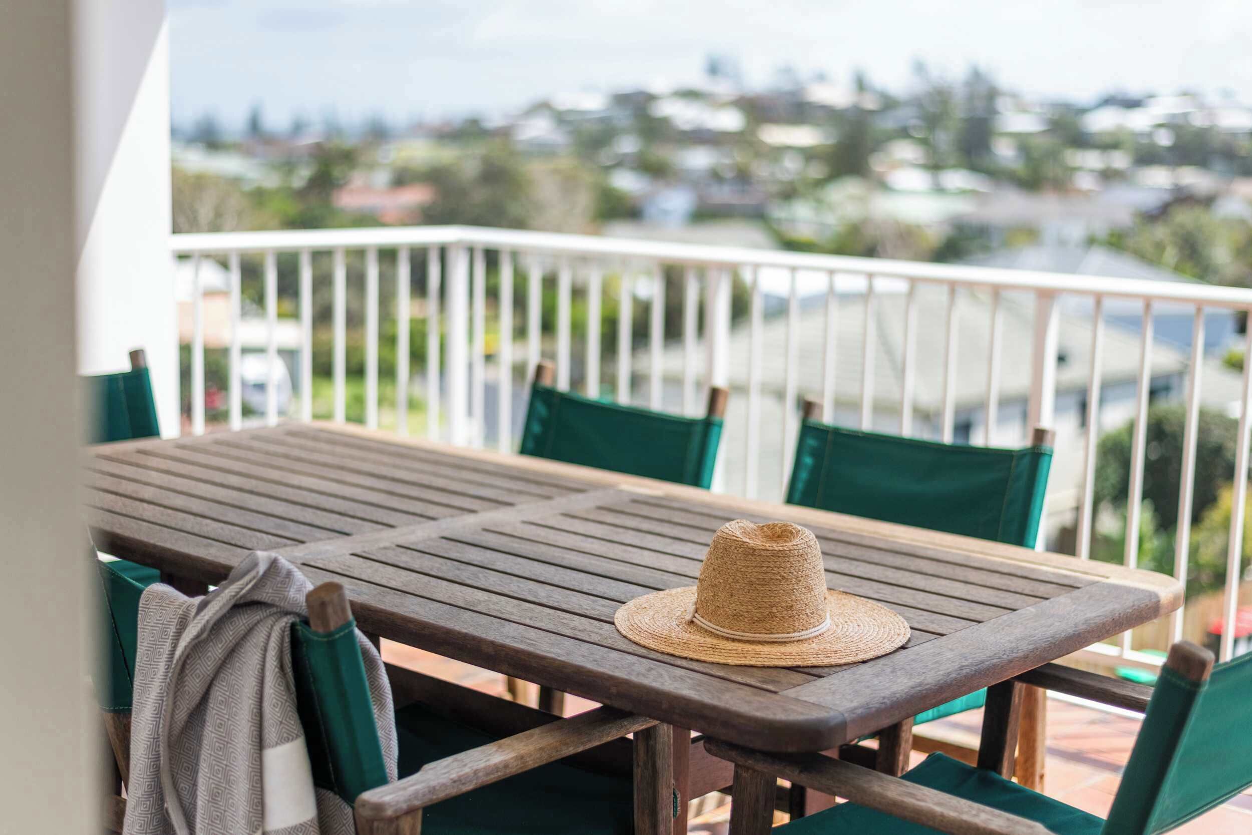Une table en bois sur laquelle est posé un chapeau de paille, entourée de chaises vertes, sur un balcon doté d’une balustrade blanche et offrant une vue sur des maisons au loin.