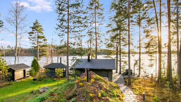 Ein Seehaus mit einem Kiesweg, der zu einem Steg führt, umgeben von hohen Bäumen und einer Wiese mit einer kleinen Gartenhütte.