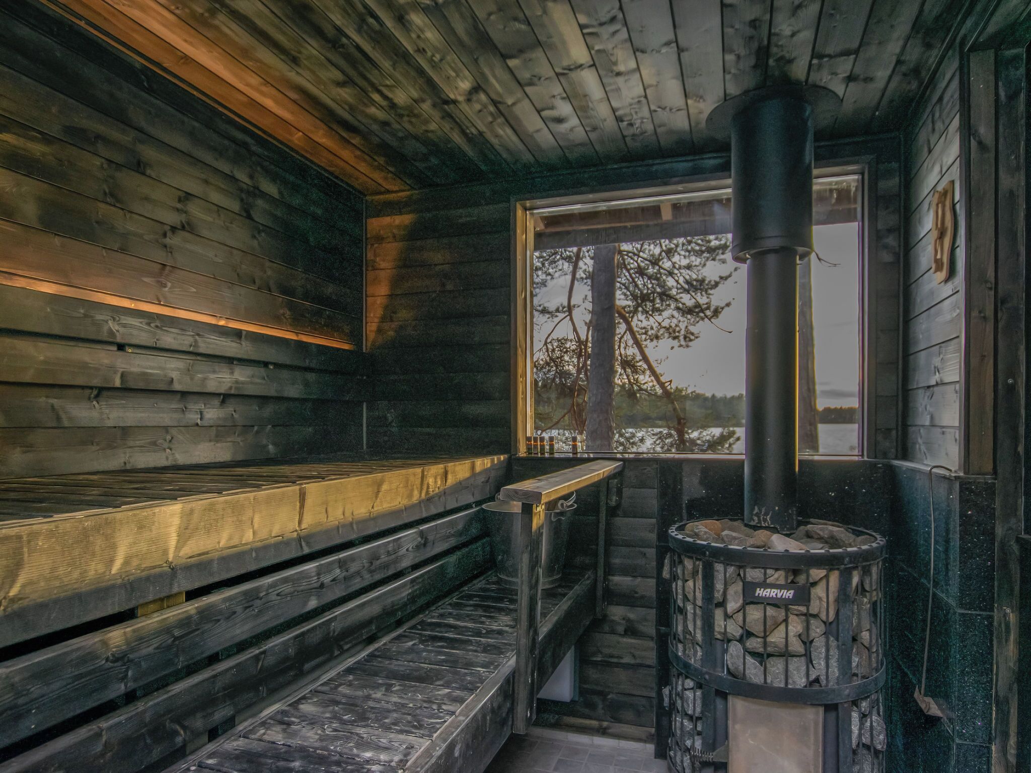A wooden cabin interior with a stone fireplace, benches, and a window overlooking a forest.