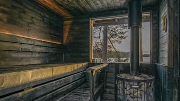 Interior de una cabaña de madera con chimenea de piedra, bancos y ventana con vista a un bosque.