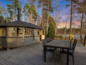 Une terrasse en bois avec une table et des chaises, une cuisine extérieure couverte et une vue sur une zone boisée.