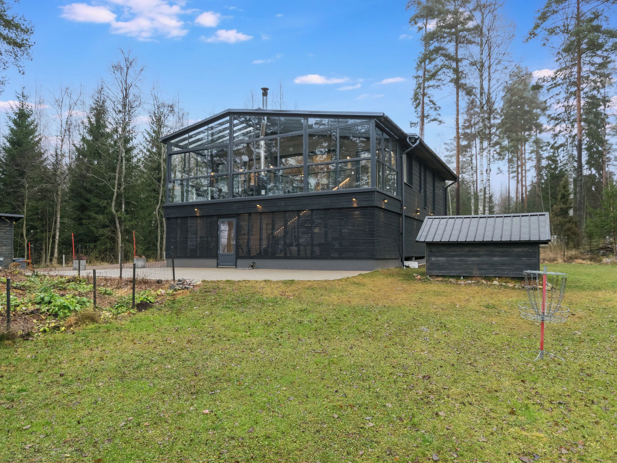 Moderni lasi- ja puurakenteinen mökki, jossa on näkyvä portaikko ja pieni, sen vieressä sijaitseva varasto.