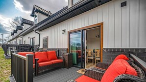En overdækket terrasse med fletningsmøbler og røde puder, en skydedør der fører til et spiseområde indendørs, samt en hvid bygning med mørk kanting.