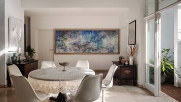 A dining area with a round table, white chairs, a large abstract painting on the wall, and a view through a glass door.