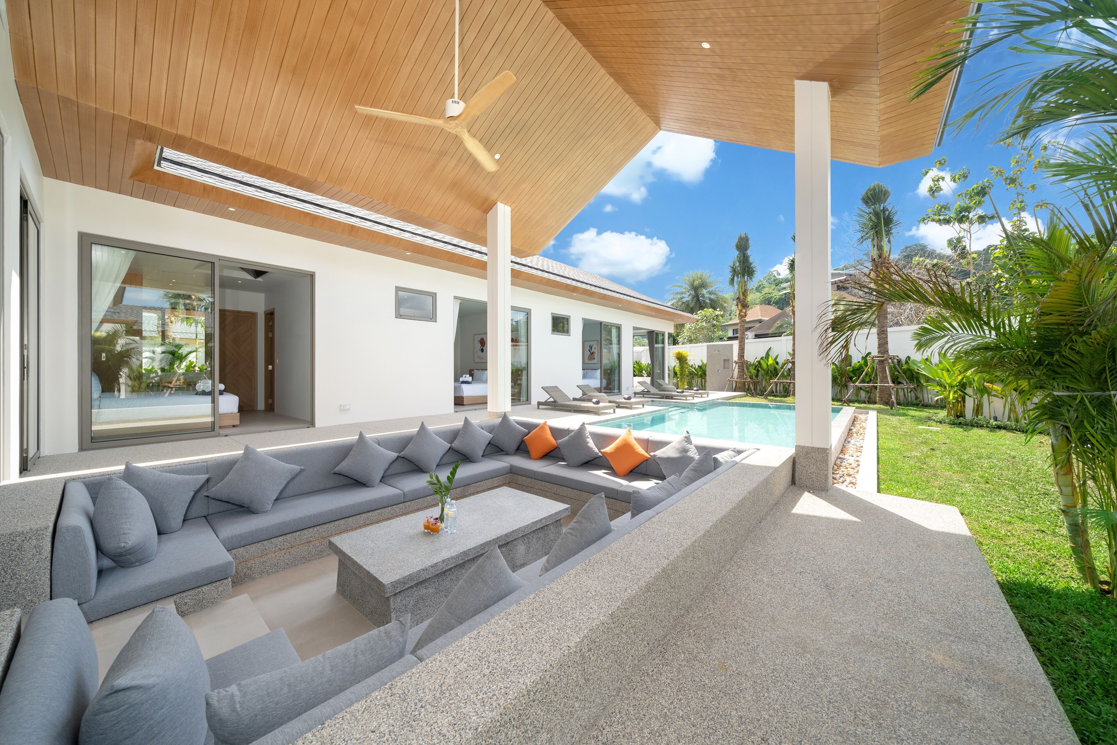 Uma área externa moderna com piscina, almofadas cinzas e laranjas, e um ventilador de teto.