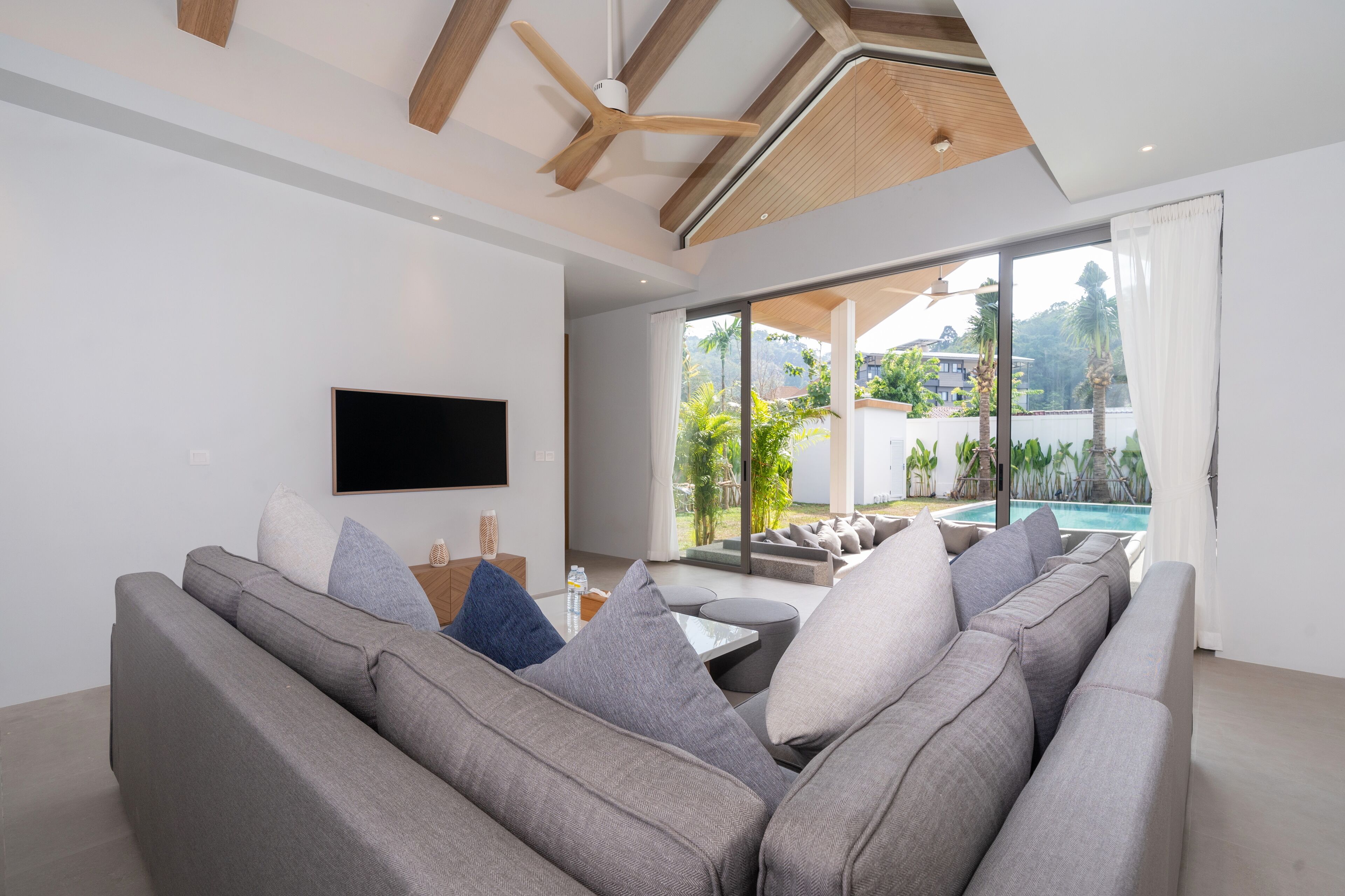 A modern living room with a grey sectional sofa, a flat-screen TV, and a large window overlooking a pool and greenery.