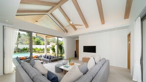 A modern living room with a large grey sofa, a coffee table, and a flat-screen TV. There is a sliding glass door leading to a pool area with outdoor seating. - Himmapana Villas - Grand Valley (Kamala)
