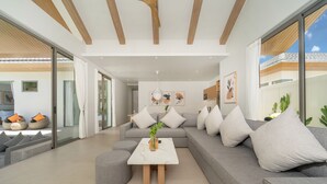 A modern living room with a grey sectional sofa, a white coffee table, and a glass door leading to an outdoor area. - Himmapana Villas - Grand Valley (Kamala)