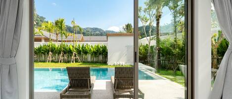 Une chambre avec vue sur une piscine, deux fauteuils en osier et une porte vitrée donnant sur un espace extérieur agrémenté de verdure et d’un bâtiment en arrière-plan.