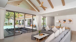 A modern living room with a large gray sectional sofa, a coffee table, and a dining area with a wooden table and chairs. - Himmapana Villas - Grand Valley (Kamala)